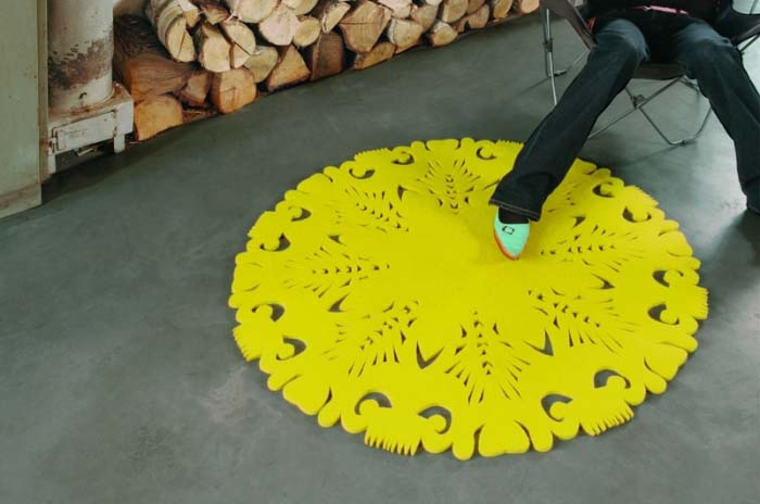 Żółty, okrągły dywanik z wycinanym wzorem na szarym betonowym podłożu, częściowo zasłonięty przez siedzącą osobę w dżinsach i turkusowym bucie, w tle sterta drewna kominkowego i fragment grzejnika.